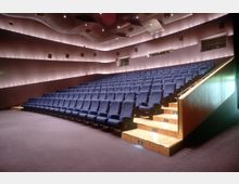 Innenraum eines modernen Auditoriums mit ansteigenden Sitzreihen aus blauen gepolsterten Stühlen. Die Wände sind sanft beleuchtet, und die Decke hat eine geschwungene Form mit eingebauten Scheinwerfern und Lautsprechern.