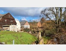 Ein ländlicher Hof mit Fachwerkgebäuden und einem größeren Holzbau, umgeben von grüner Wiese und kahlen Bäumen. Im Hintergrund ist eine historische, halb zerfallene Ziegelruine zu sehen, eingebettet in eine naturlandschaftliche Umgebung.