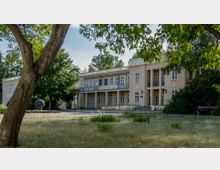 Ein zweistöckiges Gebäude mit beiger Fassade, großen Fenstern und einem Balkon, umgeben von Bäumen und einer Wiese. Im Vordergrund sind ein Baumstamm und eine Sitzbank zu sehen.