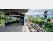 Bahnhof mit einem überdachten Bahnsteig auf der linken Seite, der von Glas- und Metallwänden begrenzt wird, sowie Gleisen auf der rechten Seite, umgeben von dichter Vegetation. Im Hintergrund sind Signalanlagen und ein Wegweiser mit der Aufschrift „Yorckstraße/Bautzener Straße“ sichtbar.