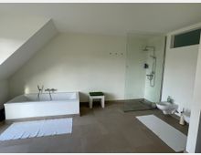 Modernes Badezimmer mit einer weißen Badewanne auf der linken Seite, einer bodengleichen Glasdusche rechts daneben und einem weißen Bidet sowie einer Wandtoilette. Der Raum hat helle Wände, eine schräge Decke und beige Fliesenboden, mit weißen Teppichen vor den Sanitäranlagen.