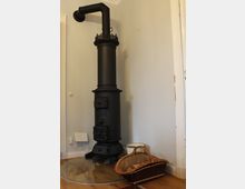 Ein schwarzer, zylindrischer Gusseisenofen mit dekorativen Verzierungen steht in einer Ecke eines Zimmers mit hellen Wänden. Daneben befinden sich ein geflochtener Korb und ein weißer Eimer auf einem Holzboden mit einer runden Glasplatte als Schutz.