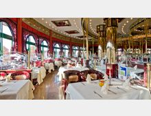 Ein elegantes Restaurant mit asiatischem Design, gekennzeichnet durch rote Säulen, große runde Fenster mit dekorativen Holzgittern und goldfarbene Elemente. Die Tische sind mit weißen Tischdecken, gefalteten Servietten und Kerzen dekoriert, während Glasabtrennungen mit gravierten Vogel- und Pflanzenmotiven den Raum strukturieren.