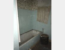 Ein veraltetes Badezimmer mit blassblauen Wandfliesen, einer verschmutzten Badewanne und einem kleinen, abgenutzten Boiler. Die Wand darüber ist mit verblasster Blumen-Tapete bedeckt, und ein heruntergekommenes Stück Stoff hängt anstelle eines Vorhangs.