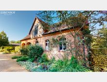 Beige Haus mit spitzem Giebel und rotem Ziegeldach, umgeben von gepflegtem Garten mit Sträuchern und Pflanzen. Links neben dem Haus steht ein kleiner Tisch mit Stühlen auf einem Schotterweg, und im Hintergrund sind Bäume und Büsche zu sehen.