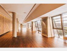 Großer, moderner Innenraum mit hellem Holzfußboden, Holzverkleidungen an einer Wand und großen schrägen Fenstern, die viel Tageslicht hereinlassen. Im Hintergrund ist eine Aussicht auf Gebäude und einen parkähnlichen Bereich zu sehen.