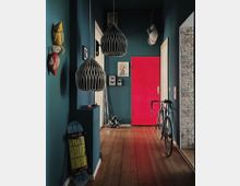 Ein modern gestalteter Flur mit dunkelgrünen Wänden, einer Holzdielung und einer auffälligen pinkfarbenen Tür. An der Wand hängen Dekorationen wie bunte Fischmasken und gerahmte Kunstwerke, während ein Rennrad und ein Skateboard den Raum ergänzen; zwei geometrisch geformte Hängelampen verleihen dem Raum zusätzlichen Charakter.