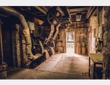 Ein alter, industrieller Maschinenraum mit sichtbaren Metallrohren, Zahnrädern und Backsteinwänden; im Hintergrund befindet sich eine offene Tür mit Blick auf eine Außentreppe und einem beleuchteten Notausgangsschild. Die Umgebung ist abgenutzt, mit Metallbehältern und einer Werkbank in einer Ecke.