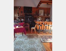 Ein Wohnzimmer oder gemütlicher Aufenthaltsraum mit Holzelementen, einer kleinen Bar mit Barhockern, bunten Teppichen auf Holzboden und dekorativ platzierten Möbeln, einschließlich eines Regals mit Figuren und einer Tischfläche mit roten Tischdecken.