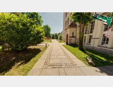 Ein gepflasterter Weg mit eingefassten Steinen führt durch eine grüne Gartenanlage mit Bäumen und Büschen. Rechts steht ein mehrstöckiges Gebäude mit hellen Fassaden und großen Fenstern, links ist ein kleiner Spielplatz sichtbar.