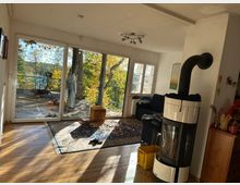 Wohnzimmer mit Holzfußboden, großem Kaminofen und bodentiefen Fenstern, die auf eine Holzterrasse mit Blick auf herbstliche Bäume führen. Im Raum stehen ein Sofa, ein Teppich und verstreute Gegenstände, bei hellem Tageslicht.