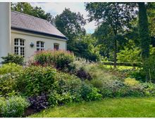 Heller Pavillon mit Schieferdach und Sprossenfenstern am Rand eines üppig bepflanzten Gartens mit Staudenbeeten und rosa Blüten. Daneben erstreckt sich eine Rasenfläche mit hohen Bäumen und dichtem Grün im Hintergrund.
