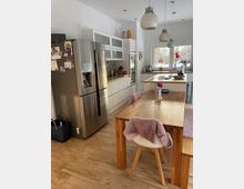 Helle, moderne Wohnküche mit weißen Einbauschränken, Edelstahl-Kühlschrank und Holzarbeitsflächen sowie großem Holzesstisch im Vordergrund. Über einer Kücheninsel und dem Tisch hängen Pendelleuchten, und ein Fenster an der Rückwand bringt Tageslicht in den Raum.