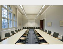 Konferenzraum mit zwei langen, weißen Tischen, die parallel zueinander aufgestellt sind, umgeben von gepolsterten Stühlen mit Armlehnen. Der Raum hat hohe Decken, große Fenster auf der linken Seite, eine weiße Projektionsfläche an der Stirnwand und minimalistische Wanddekorationen.