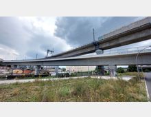 Betonbrücken in einer städtischen Umgebung, mit mehreren Ebenen, die Straßen- und Schienenverkehr tragen. Im Vordergrund eine bewachsene Grünfläche und ein Gehweg, im Hintergrund Gebäude und ein bewölkter Himmel.