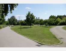 Weitläufiger Park mit grünen Rasenflächen, vereinzelten Bäumen und gebogenen gepflasterten Wegen. Im Hintergrund sind moderne Gebäude und eine Industrieanlage mit zwei Schornsteinen zu sehen.