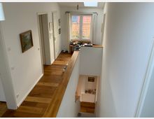 Flur in einem Wohnhaus mit Holzboden, weißen Wänden und mehreren Türen zu angrenzenden Räumen; ein Fenster mit Blick auf ein Ziegeldach befindet sich am Ende des Flurs, Treppen führen nach unten.