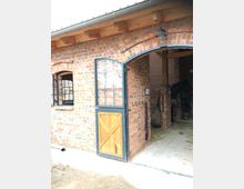 Backsteinbau mit offen stehender Stalltür aus Holz und Metall; im Inneren des Stalls sind Heu und Haken an der Wand sichtbar, ein dunkel angedeutetes Pferd steht im Hintergrund.