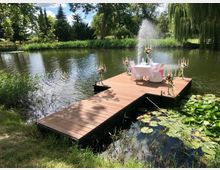 Holzsteg über einem Teich in einer grünen Parklandschaft, dekoriert mit Blumen, Kerzenleuchtern und einem Tisch für zwei Personen, der mit weißer Tischdecke gedeckt ist; im Hintergrund befindet sich eine sprudelnde Wasserfontäne und dichter Baumbestand.