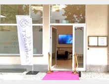 Eingang einer Zahnarztpraxis mit großen Glasfenstern, einem offenen Glastürbereich und einem pinkfarbenen Teppich, der zum Eingang führt. Links steht eine Fahne mit der Beschriftung "Zahnarzt", rechts ein Aushang mit Informationen. Im Inneren sind ein Fernseher sowie Sitzmöbel sichtbar.