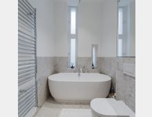 Modernes Badezimmer mit einer freistehenden weißen Badewanne, beigefarbenen Wandfliesen im unteren Bereich, einem wandhängenden WC und einem verchromten Handtuchheizkörper. Zwei schmale, hohe Fenster lassen Tageslicht herein.