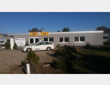 Ein einstöckiges graues Gebäude mit mehreren Fenstern und gelben Schildern mit der Aufschrift „Ankauf Verkauf“ und „Service Center“. Davor steht ein weißer Kombi auf einem unbefestigten Parkplatz, umgeben von vereinzelten Büschen und kleinen Grünflächen.