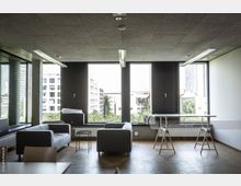 Modernes Büro mit großen Fenstern, die einen Blick auf städtische Gebäude und grüne Baumkronen bieten. Im Raum stehen graue Sofas, ein runder Glastisch und minimalistische Holztische mit Böcken auf Parkettboden.