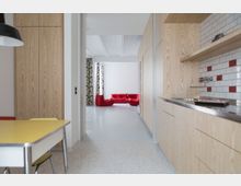 Moderne Wohnküche mit hellen Holzmöbeln, einer Arbeitsplatte aus Edelstahl und einer Rückwand mit weißen und roten Fliesen. Im Hintergrund ein offener Wohnbereich mit roten Polstermöbeln und bedruckten Vorhängen.