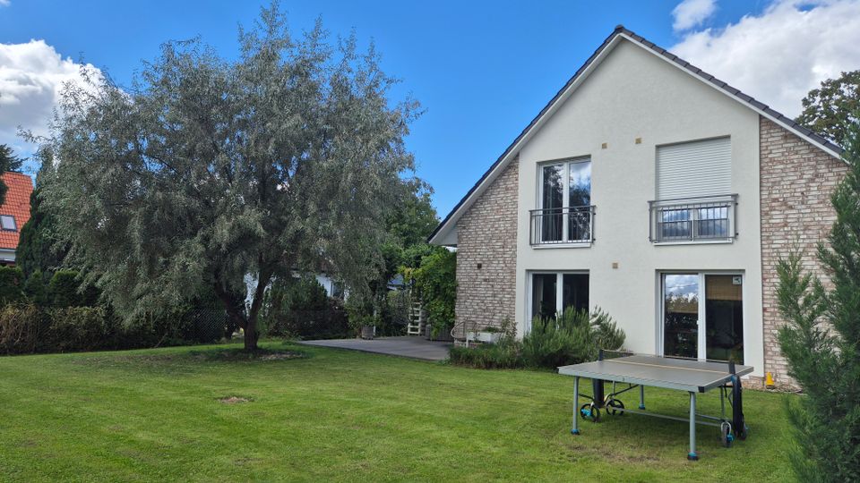 Ein Einfamilienhaus mit heller Fassade und Teilen aus Klinkerstein, zwei Balkonen und großen Fenstern. Im Vordergrund ein gepflegter Garten mit einem großen Baum und einer Tischtennisplatte, umgeben von Büschen und Rasen.