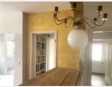 Raum mit gelber Wand, einer hölzernen Tischplatte und einem Kronleuchter mit Messingarmen und einer weißen Glaskugel. Im Hintergrund sind ein Durchgang mit Bücherregalen und ein Fenster in einer Küche zu sehen.