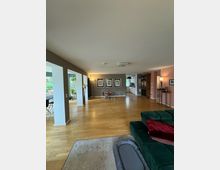 Großes, modern eingerichtetes Wohnzimmer mit Holzboden und beigen Wänden. Der Raum enthält eine grüne Couch, einen Teppich, eine Wand mit Bildern sowie einen Essbereich und eine offene Küchenecke im Hintergrund. Große Fenster bieten Blick ins Freie und lassen viel Tageslicht herein.