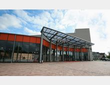 Außenansicht eines modernen Gebäudes mit einer Glasfassade, orangefarbenen Paneelen und einem weit auskragenden Glasvordach, das von Metallträgern gestützt wird. Im Vordergrund ist ein großer gepflasterter Platz unter freiem Himmel zu sehen.