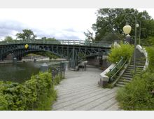 Historische Stahlbrücke über einen ruhigen Fluss, flankiert von einer gepflasterten Uferpromenade mit Laternen und Treppenaufgang, umgeben von üppigem Grün. Im Hintergrund sind Bäume und Geländer der Brücke zu sehen.