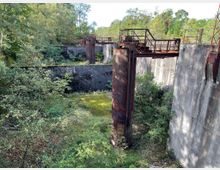 Verlassene Industrieanlage mit einer massiven Betonmauer, rostigen Metallstrukturen und überwucherter Vegetation; im Hintergrund dichter Wald.