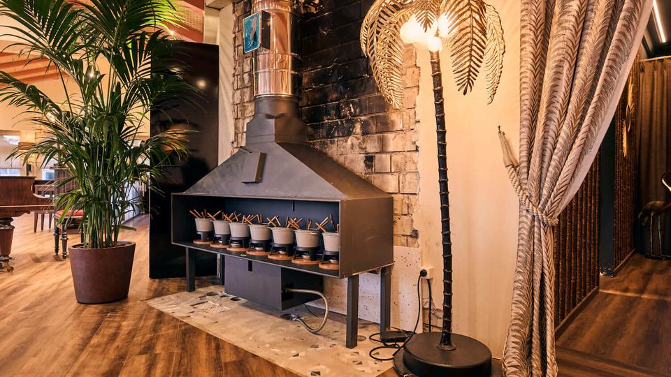Warmly lit restaurant interior with wooden floors, a large black metal fireplace hood and chimney against a dark brick wall, and a potted palm beside it. A palm-shaped floor lamp and patterned curtain partition define a cozy corner of the space.