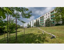 Wohngebiet mit vierstöckigen Mehrfamilienhäusern, weißen Fassaden und grünen Balkonen. Im Vordergrund eine Wiese mit einem kleinen Spielplatz, einschließlich einer Wippe und kletterähnlichen Metallstrukturen, umgeben von Bäumen und Sträuchern.