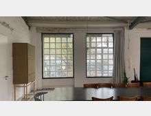 Schlichter Innenraum mit zwei großen, gitterförmigen Fensterfronten, die Tageslicht hereinlassen. Vor den Fenstern steht ein langer Konferenztisch mit Stühlen; links ein hoher Schrank und rechts Vorhänge sowie einige Pflanzen vor einer dunklen Tür.