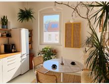 Helle, minimalistisch eingerichtete Küche mit einer weißen Kühl-Gefrierkombination, hölzernen Arbeitsflächen, offenen Regalen und einer hängenden Zimmerpflanze. Ein runder Esstisch mit Rattanstühlen steht vor einem Fenster, das den Blick auf ein Gebäude mit roten Ziegeldächern bietet; mehrere Zimmerpflanzen und ein gelbes Wandpaneel schmücken den Raum.