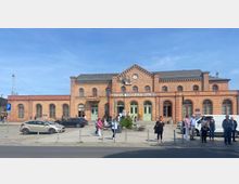 Bahnhofsgebäude in Königs Wusterhausen aus rotem Ziegelstein mit Rundbogenfenstern, grünen Eingangstüren und einer zentralen Uhr an der Fassade. Davor ein gepflasterter Vorplatz mit parkenden Autos, Fußgängern und wartenden Personen.