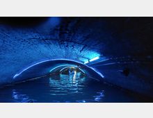 Ein unterirdischer Backsteintunnel mit einem Wasserkanal, beleuchtet durch blaue Neonlichter, in dem ein kleines Boot zu sehen ist. Rohre verlaufen entlang der Wände und der Decke des Tunnels.