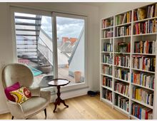 Wohnzimmer mit einem großen Fenster, das auf eine Dachterrasse mit Metalltreppe und Pflanzentöpfen blickt. Innen steht ein beiger Sessel mit bunten Kissen neben einem kleinen Holztisch, und eine weiße Bücherwand voller Bücher bedeckt die rechte Seite des Raumes.