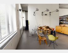 Ein heller Arbeitsraum mit großen Fenstern auf der linken Seite, durch die Tageslicht einfällt. Im Raum stehen verschiedene moderne Stühle, Arbeitstische und kleine mechanische Geräte; an den Wänden sind Lampen mit schwenkbaren Armen angebracht.