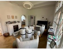 Wohnzimmer mit hellen Wänden und Parkettboden, ausgestattet mit einem weißen Kamin, einem schwarzen Klavier und Sesseln. Große Fenster mit bunten Bleiglasverzierungen lassen Tageslicht hinein; ein kleiner Tisch und Bücherregale ergänzen die Einrichtung.