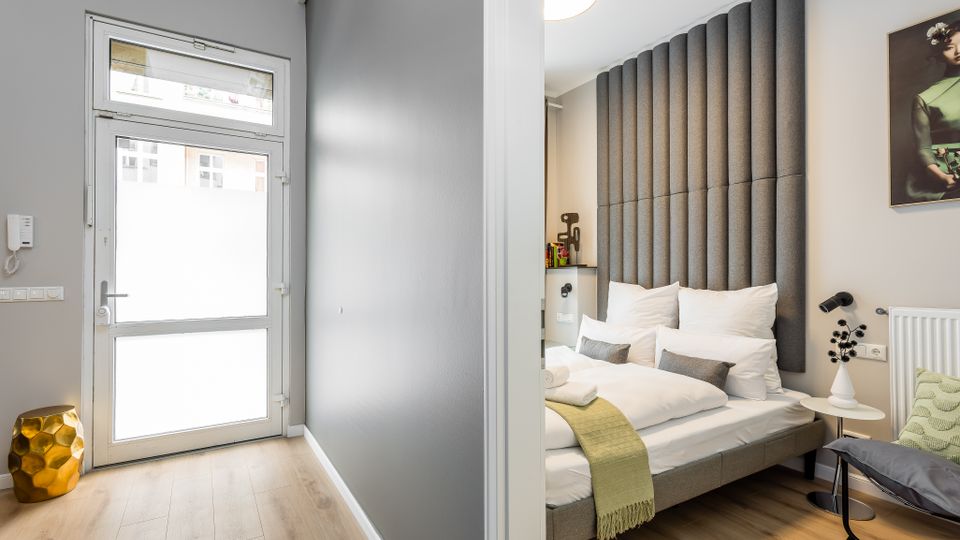 Blick in ein modernes Schlafzimmer mit Doppelbett, hoher gepolsterter Wandverkleidung als Kopfteil und heller Holzoptik am Boden. Links führt ein schmaler Flur zu einer Glastür, rechts stehen ein kleiner runder Beistelltisch mit Lampe und ein Heizkörper an der Wand.