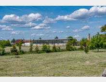 Eine industrielle Halle mit großen Fenstern steht inmitten einer ländlichen Landschaft, umgeben von Wiesen, Bäumen und einem Maschendrahtzaun. Im Hintergrund sind Windräder, kleinere Gebäude und ein hoher Schornstein zu sehen, unter einem blauen Himmel mit Wolken.