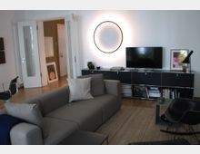 Modernes Wohnzimmer mit einem grauen Sofa, einem Fernseher auf einem schlichten schwarzen Regal und einer großen, beleuchteten Wanddekoration in Form eines Kreises. Der Raum hat helle Wände, einen Holzboden und Zugang zu einem offenen Flur durch weiße Doppeltüren.