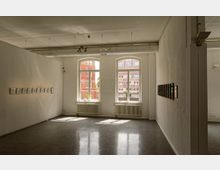 Minimalistischer Galerieraum mit weißen Wänden, Hängeschienenbeleuchtung und zwei großen, bogenförmigen Fenstern, die Tageslicht hereinlassen. An beiden Wänden befinden sich kleine Kunstwerke in Rahmen, während der Boden aus poliertem, dunklem Material besteht.