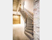 Innenansicht eines Treppenhauses mit breiter Betontreppe und schwarzem Metallgeländer mit Holzhandlauf, die in einem Bogen nach oben führt. Helle Wände, eine sichtbare Backsteinwand im Vordergrund und doppelflügelige Holztüren im Erdgeschoss prägen den Raum.