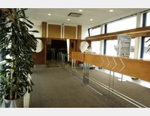 Innenraum eines Empfangs- oder Wartebereichs mit hölzernen Absperrungen und Schildern. Große Fenster lassen Tageslicht herein, und im Hintergrund befinden sich Durchgänge mit runden Fensteröffnungen in den Türen. Links steht eine grüne Pflanze in einem Topf.