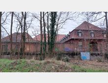 Backstein-Gebäude mit roten Ziegeldächern, teilweise verwittert, umgeben von kahlen Bäumen und einem Zaun. Im Vordergrund ist verwilderte Vegetation zu sehen, und das Gebäude trägt Graffiti an den Wänden.